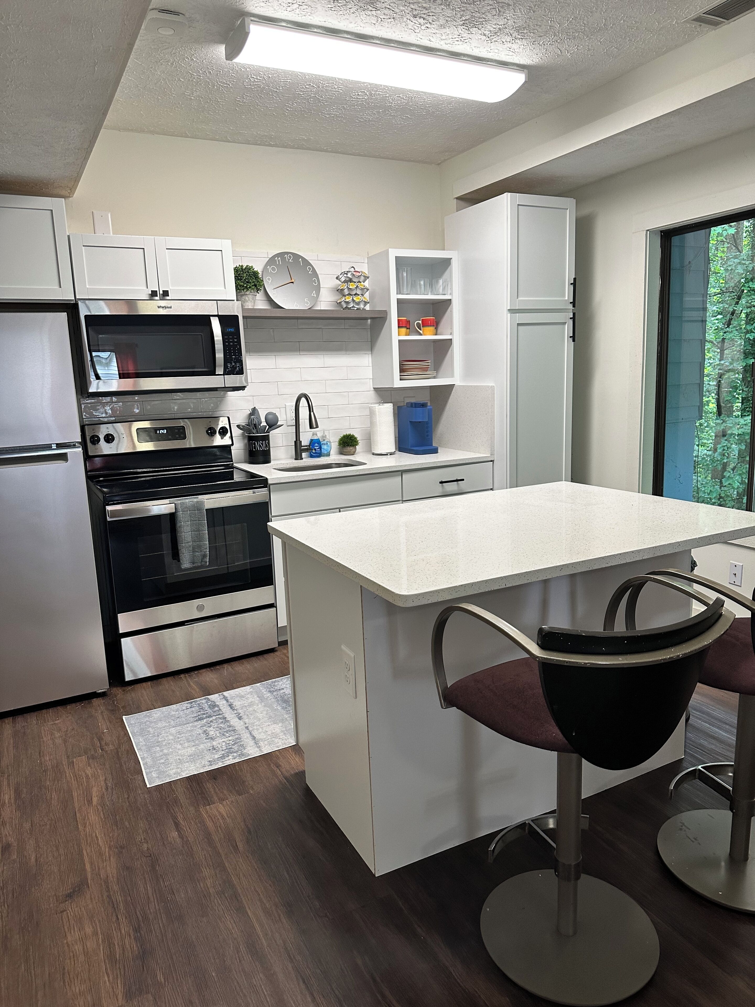 A modern kitchen with white cabinetry, a stainless steel refrigerator, a microwave, an oven, and a sink. There is a bar stool with a black seat and a white countertop.