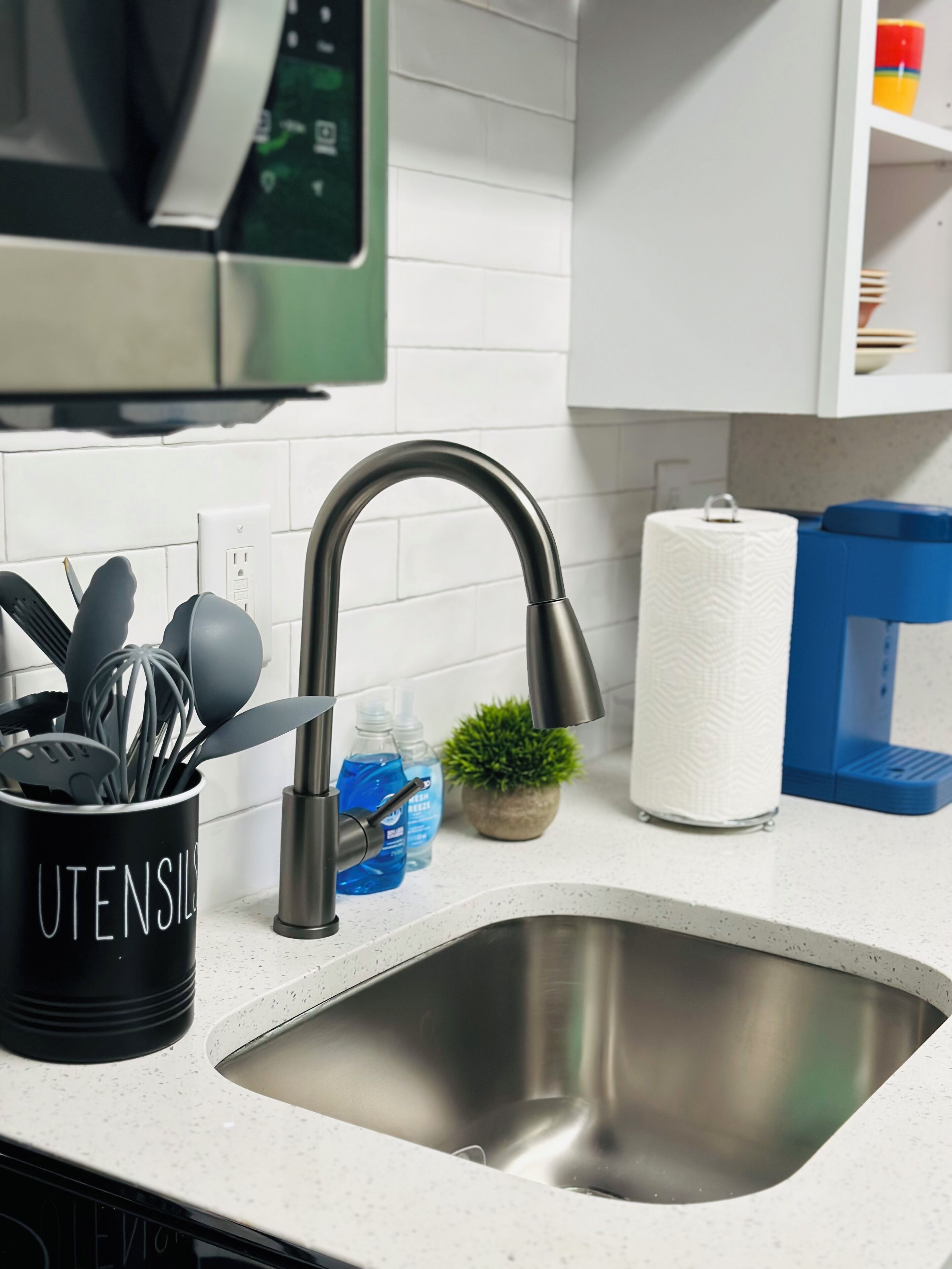 A kitchen with a stainless steel sink, a utensil holder, a paper towel dispenser, and a microwave above the sink.