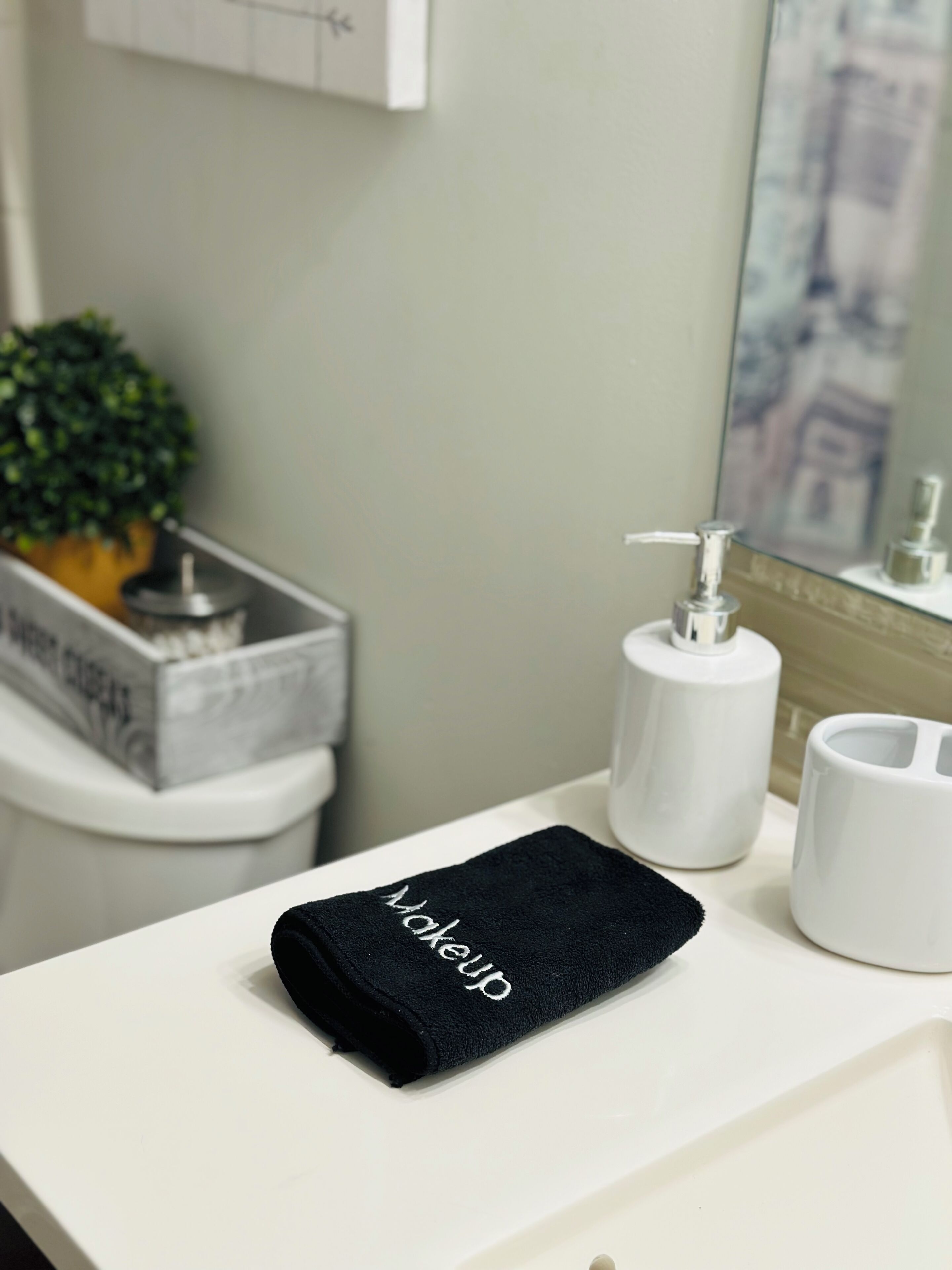 A bathroom with a white sink, a black towel with the word "Wakeup" on it, a white soap dispenser, and a potted plant.