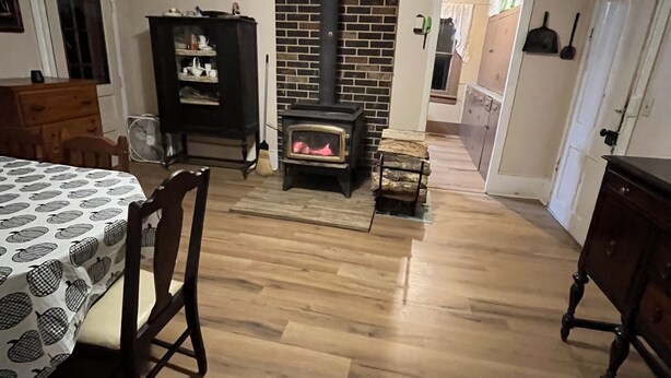 A room with a brick fireplace, wooden flooring, a dining table with a patterned tablecloth, and a dark wooden cabinet. - Charming Farmhouse on a working farm. (Pittsfield)