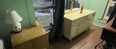 A room with a wooden dresser, a lamp, a window with curtains, and a mirror.