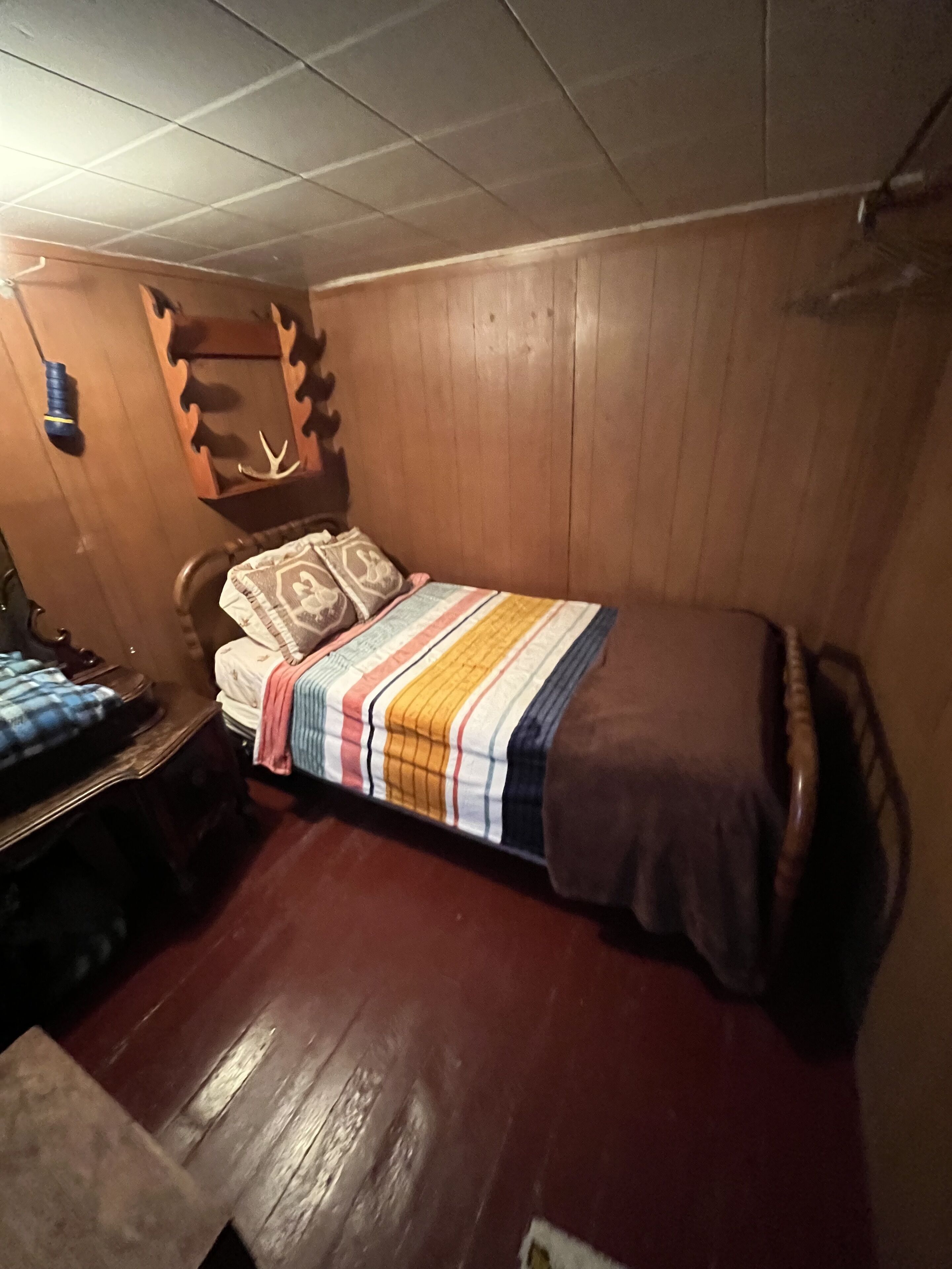 Een kleine, gezellige kamer met een houten vloer en wanden, een bed met een gestreepte deken, een houten hoofdbord en een klein tafeltje met een stoel.