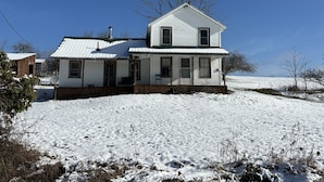 A two-story white house with a green roof, surrounded by snow-covered ground and trees. - Charming Farmhouse on a working farm. (Pittsfield)