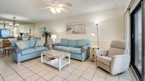 A living room with a blue sofa, a beige armchair, a wooden coffee table, a ceiling fan, and a large window. - Moondrifter #305 2 BR - Free Beach Chairs! (Panama City Beach)