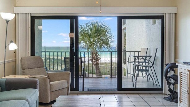 A room with a ceiling fan, a sofa, a dining table with chairs, and a view of the ocean through a sliding glass door. - Moondrifter #305 2 BR - Free Beach Chairs! (Panama City Beach)