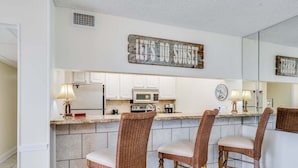 A kitchen with a breakfast bar, wicker chairs, a microwave, and a clock. - Moondrifter #305 2 BR - Free Beach Chairs! (Panama City Beach)