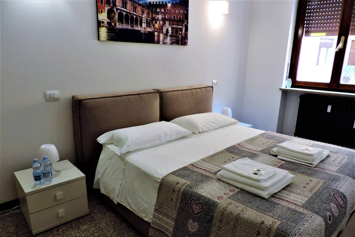 Hotelkamer met een bed, nachtkastje, waterfles, lamp en raam met jaloezieën.