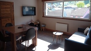 Una sala de estar acogedora con techo de madera, un ventanal con vistas a casas, una mesa de comedor con sillas, un televisor y un taburete pequeño.