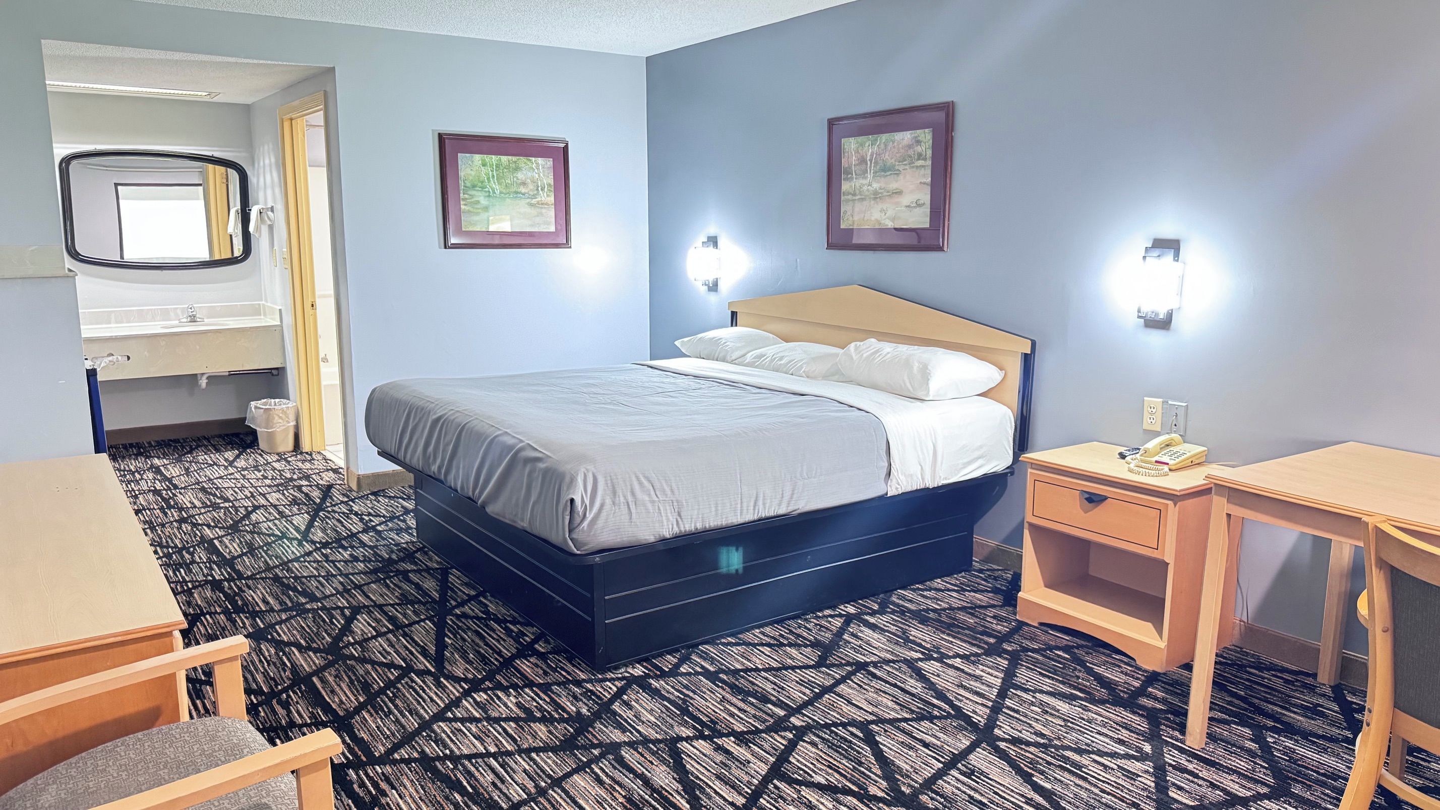 A hotel room with a bed, bedside tables, a desk, and a bathroom with a mirror and sink.