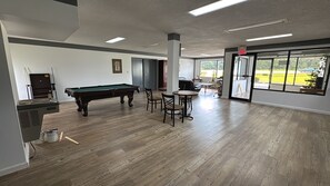 A pool table with chairs, a billiards rack, and a pool cue in a room with wooden flooring and large windows. - Westen Motel (Scooba)
