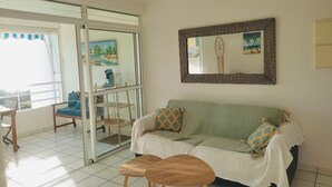 Una sala de estar con un sofá, dos taburetes de madera, una mesa de vidrio y una silla. Se observa un espejo grande en la pared y un cuadro sobre el sofá.