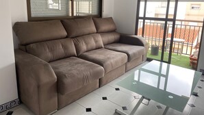 Un salon moderne avec un canapé d’angle marron, une table basse en verre et une vue sur un jardin verdoyant, visible à travers de grandes fenêtres.