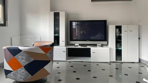 Un salon moderne doté d’un téléviseur à écran plat, de meubles blancs et d’un pouf à motifs.
