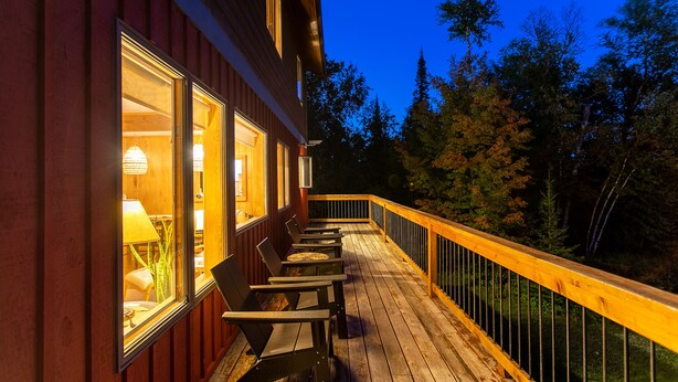 A wooden deck with outdoor seating and a railing, overlooking a forested area.