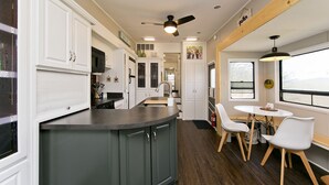 A modern kitchen with a central island, white cabinetry, and a dining area with a table and chairs. - Large Glamper w/ Hot Tub and full bathroom-sleep 6 (Capon Bridge)