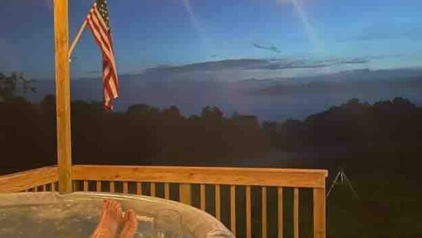 A hot tub on a wooden deck with an American flag, string lights, and a view of the landscape. - Large Glamper w/ Hot Tub and full bathroom-sleep 6 (Capon Bridge)