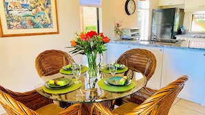 Un coin repas avec une table en verre entourée de chaises en osier, un vase rempli de fleurs et, au fond, une cuisine.