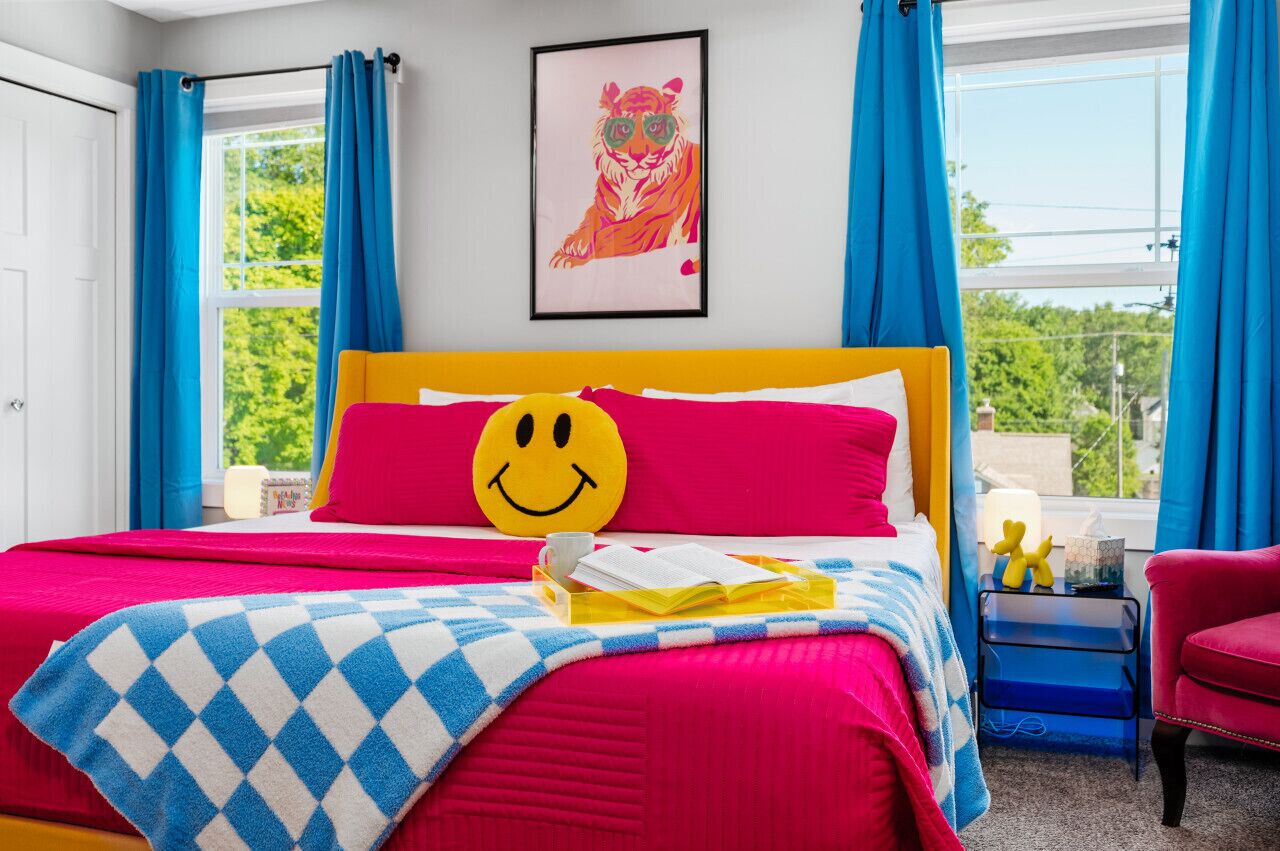 A brightly colored bedroom with a bed, a smiley face pillow, a framed tiger artwork, and a window with curtains.
