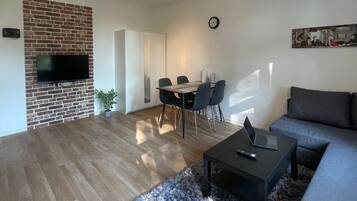 Een moderne woonkamer met een metselwerk open haard, een flatscreen televisie, een eethoek met een tafel en stoelen, een grijze bank en een pluizig vloerkleed.