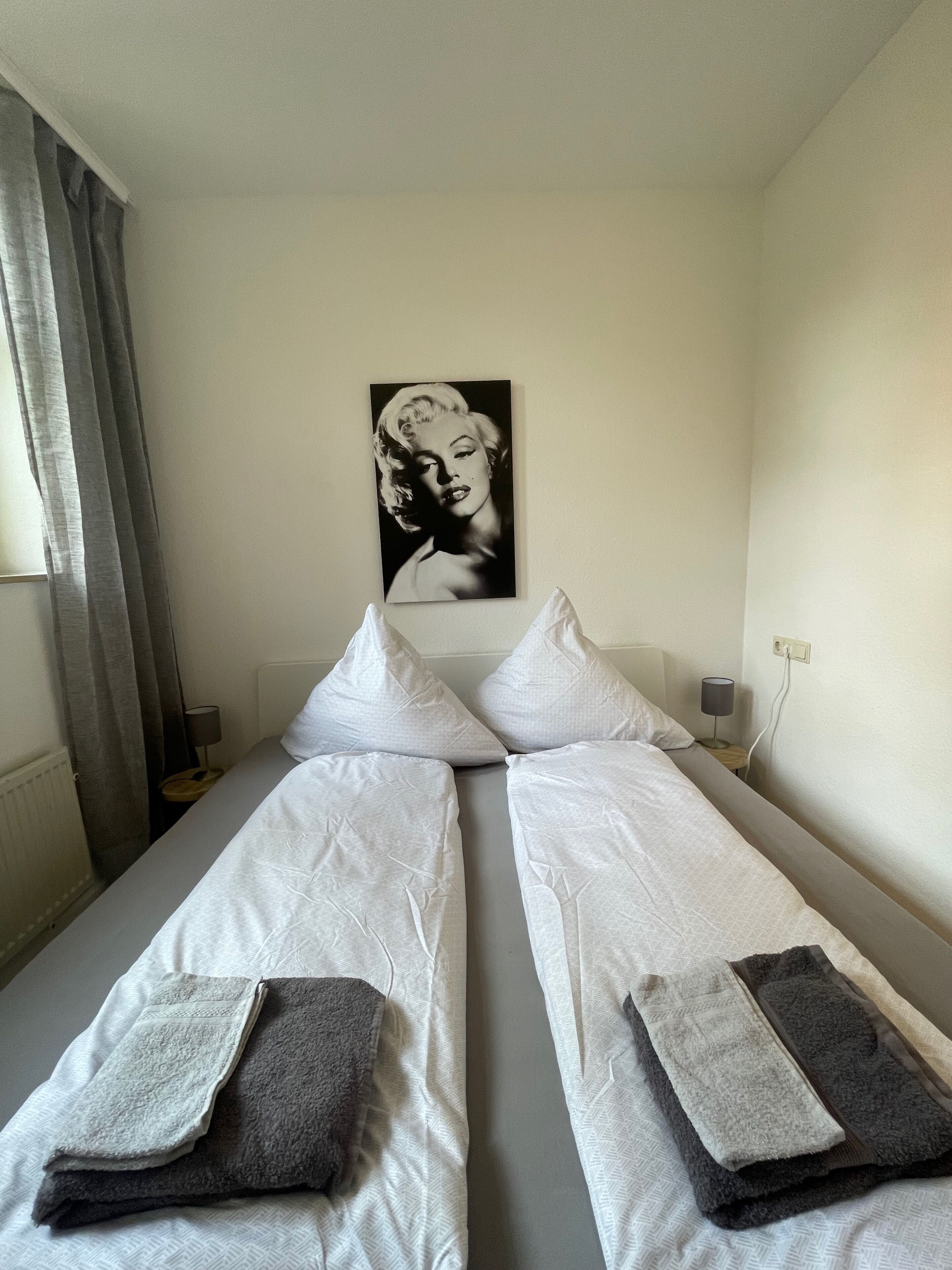 Ett hotellrum med två separata sängar, en grå sänggavel, en vit vägg, ett svartvitt porträtt och ett fönster med grå gardiner.