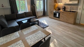 A modern living room with a sofa, dining table, and chairs. There is a kitchen area with cabinets and a stove. Large windows allow natural light to enter.