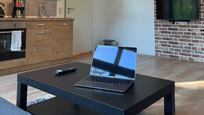 A modern kitchen with white cabinets, a coffee machine, and a microwave. There's a laptop on a table with a remote control beside it. A TV is mounted on a brick wall.