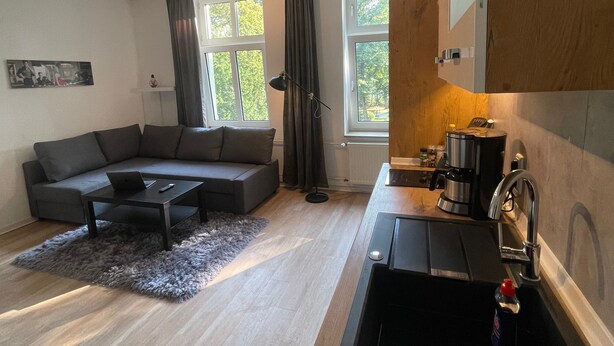 A modern living room with a grey sectional sofa, a small coffee table, and a black floor lamp. There is a kitchen area with a sink, a stove, and a countertop. Large windows allow natural light to enter the room.
