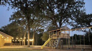 A playground with a slide and swings, surrounded by trees and a building with string lights. - Secluded Whoop Acre Ranch SLEEPS 25-LOADED with entertainment (Bryan)
