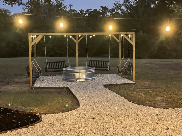 A swing set with a metal bench in the center, surrounded by pebbles, under string lights.