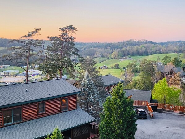 Eine Hütte mit Veranda, ein Auto auf der Auffahrt und Blick auf eine hügelige Landschaft mit Bäumen und einem Gebäude in der Ferne.