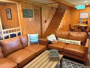 Une piÚce chaleureuse avec un canapé en cuir marron, un escalier en bois et un coin repas comprenant une table et des chaises.