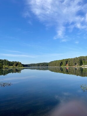 A calm lake with clear reflections, surrounded by dense forest and a few buildings. - Cozy Vermont Lake Lodge with Kayaks Fishing & Campfire Fun (Ryegate)