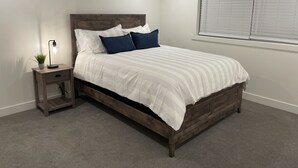 Un dormitorio con cama de madera, ropa de cama blanca, una mesita de noche con lámpara y una ventana con persianas.