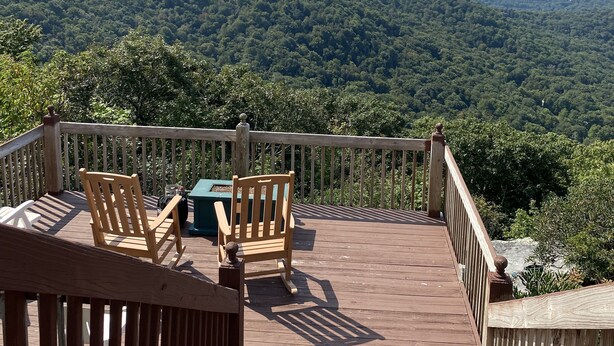 Terraza de madera con dos sillas y una mesa, con vista a una cadena montañosa boscosa.