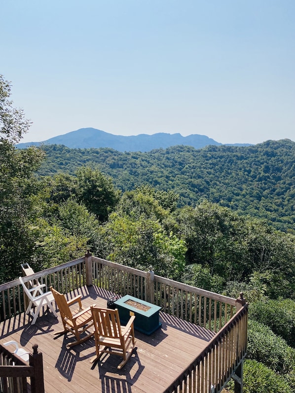A wooden deck with outdoor furniture, including chairs and a table, overlooking a forested mountain range.