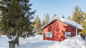 Une cabane en bois rouge, dont le toit est recouvert de neige, entourée d’arbres et d’une pile de neige.