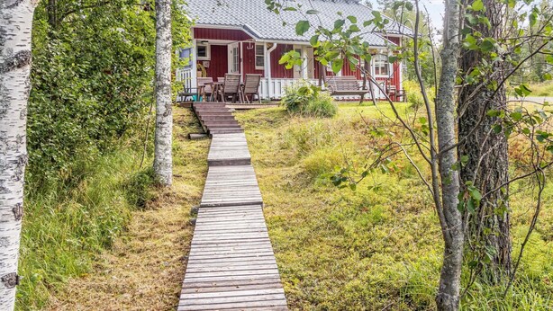 Un sentier en bois mène à une maison rouge dotée d’un porche, qui est entourée de verdure et d’arbres.