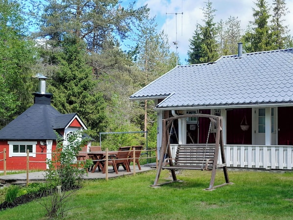 Une cabane rustique dotée d’une cheminée, d’une balançoire en bois et d’un coin repas avec une table et des chaises.