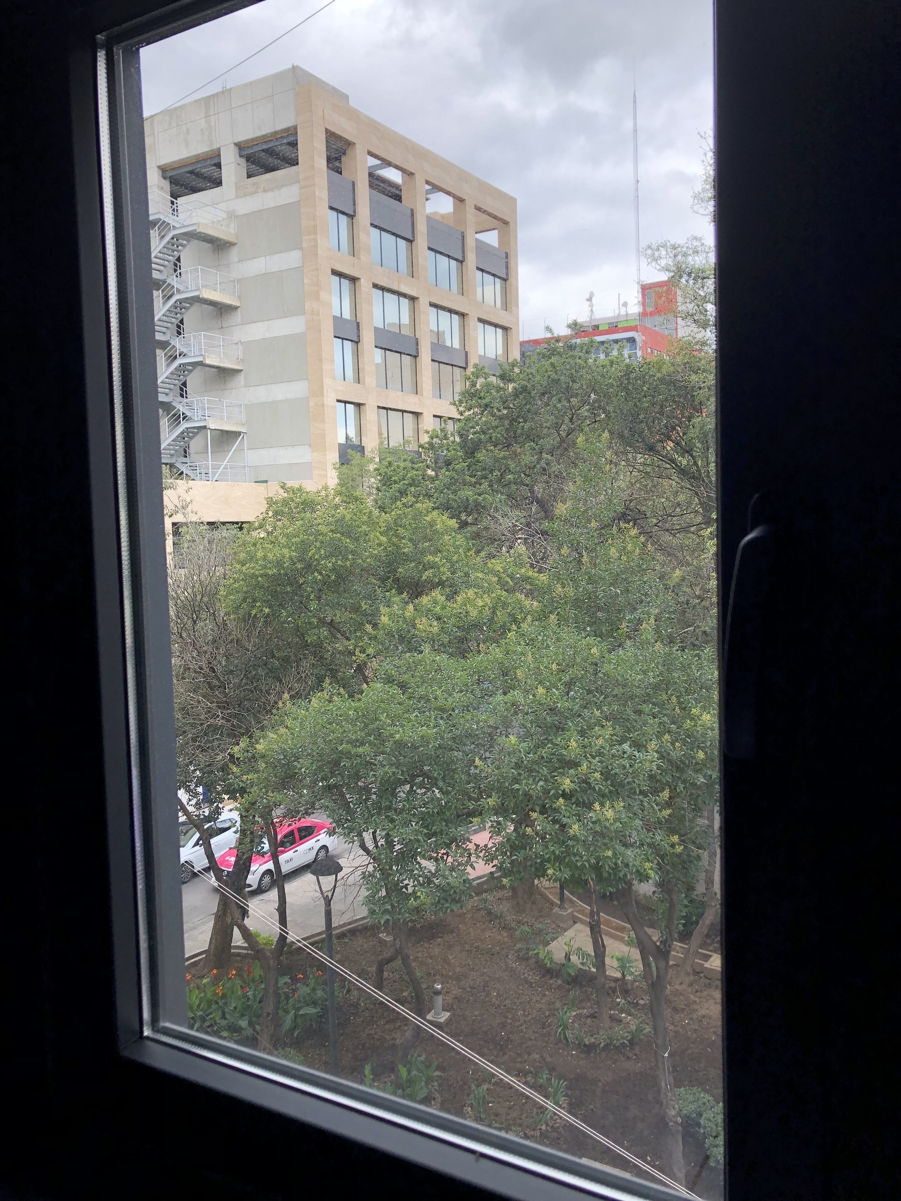 Vista a través de una ventana que muestra un edificio de varios pisos, una calle arbolada y coches estacionados.