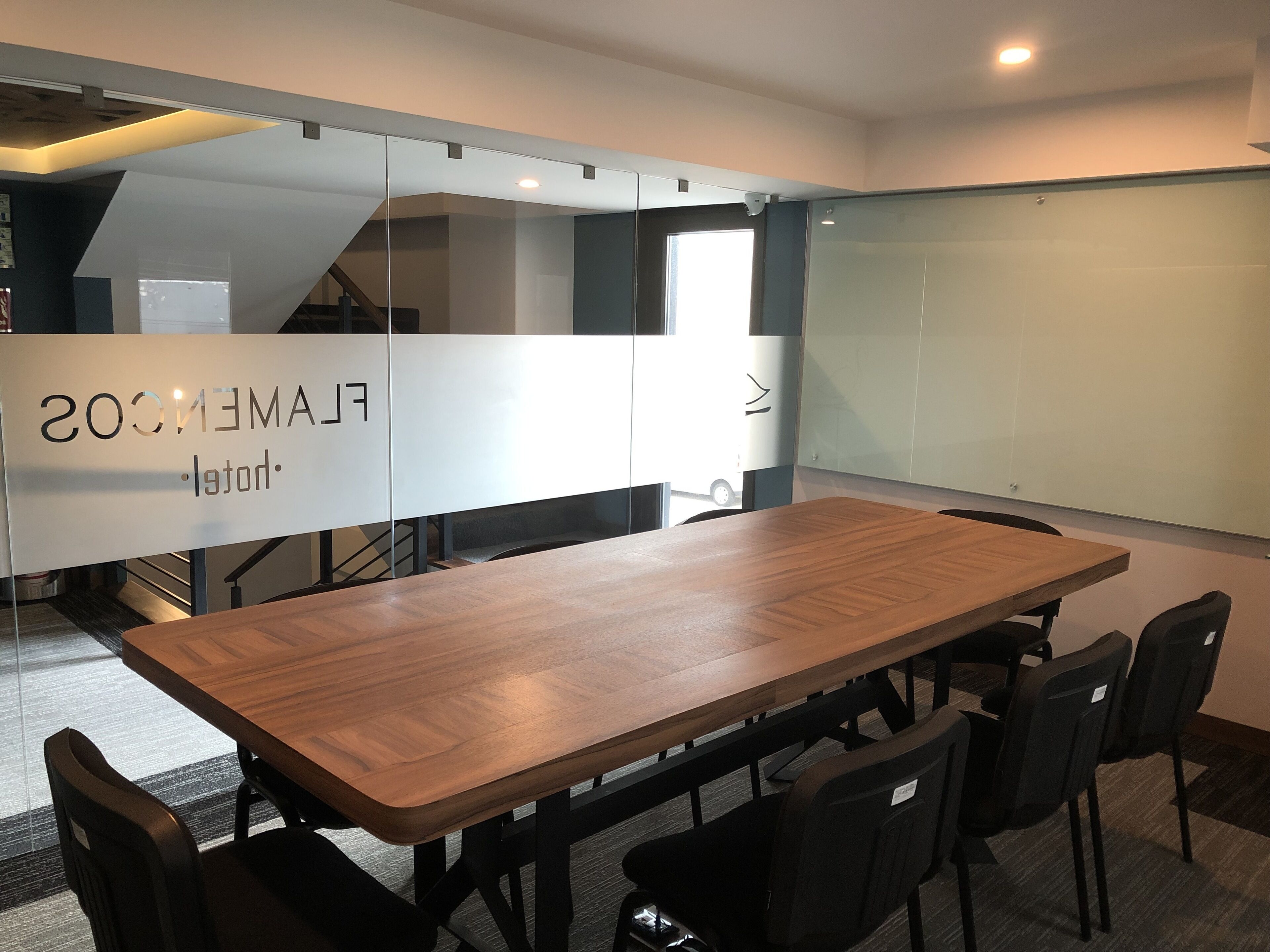 Sala de conferência com uma longa mesa de madeira, cadeiras pretas, quadro branco e um painel de vidro com texto.