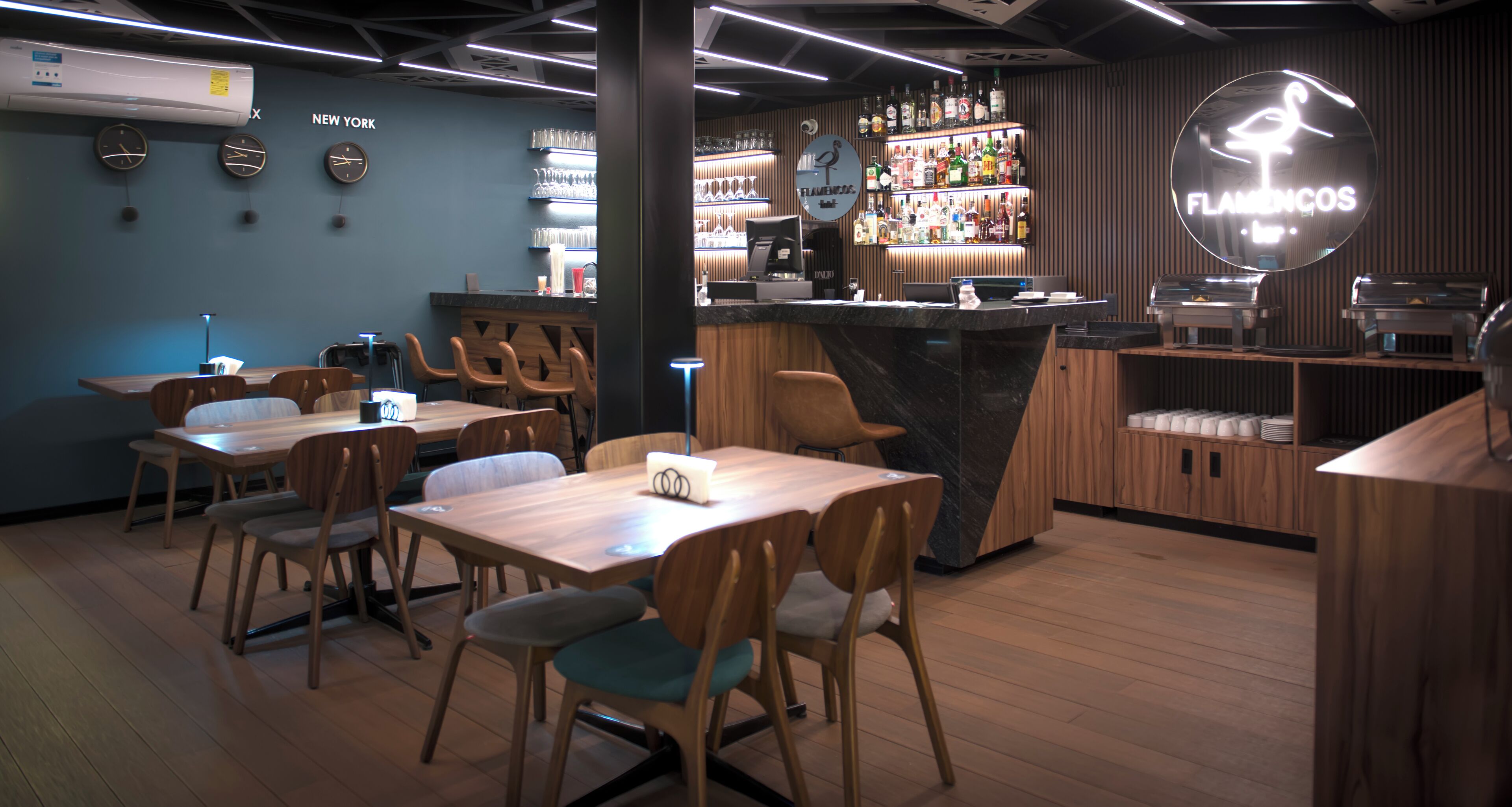 Un área de bar moderna con mobiliario de madera, una barra con un letrero de neón y una pared decorada con relojes y un cartel que dice 'NEW YORK'.