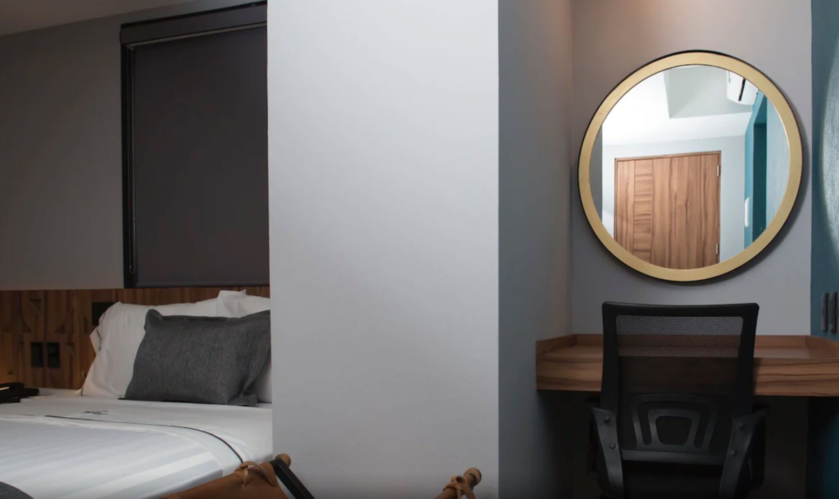 A bedroom with a bed, a desk, and a chair. There is a round mirror on the wall.