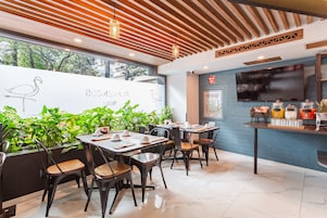 A modern restaurant interior with wooden ceiling beams, black metal chairs, and a large window overlooking greenery.