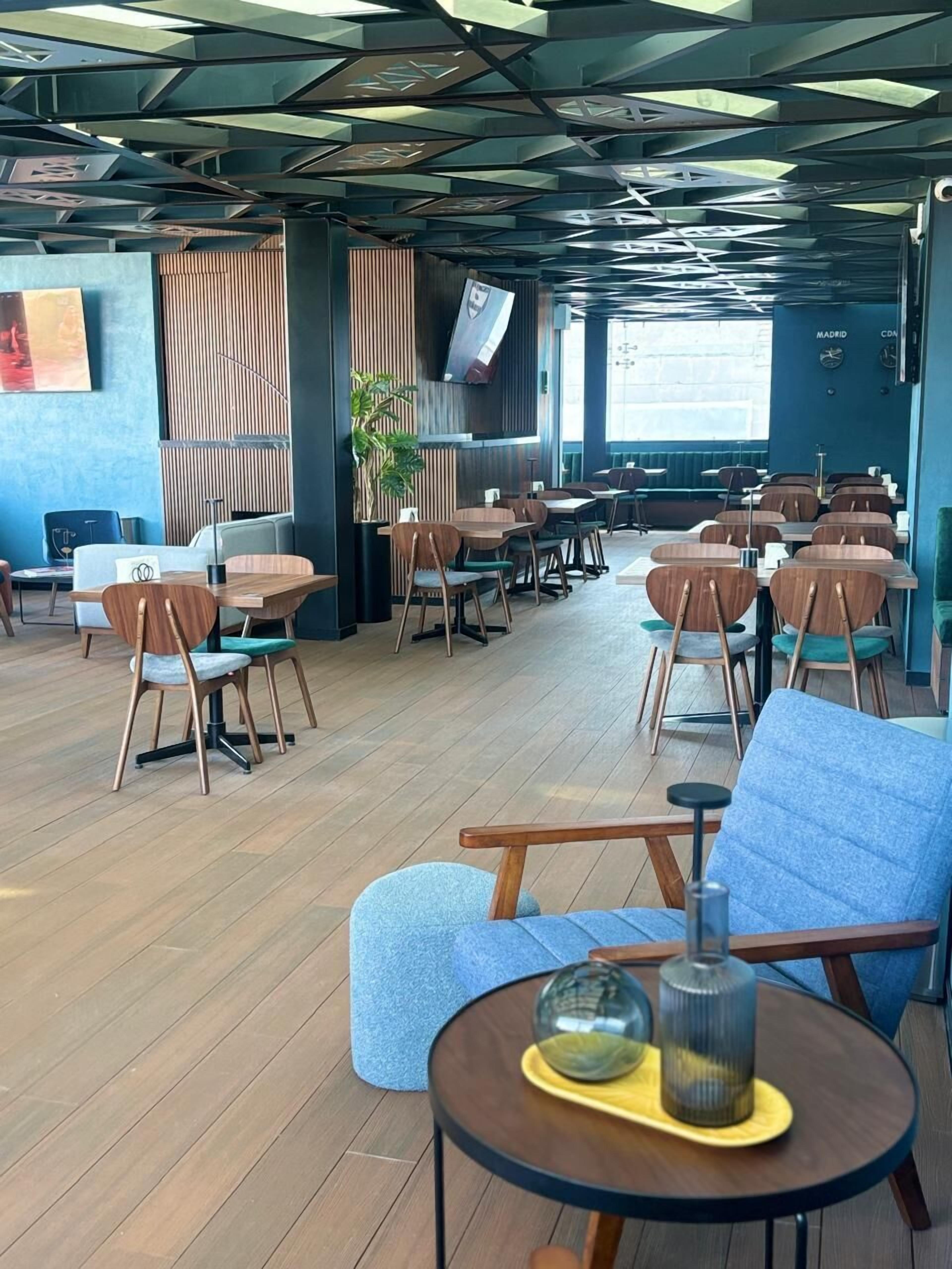 A modern restaurant interior with wooden tables and chairs, a large TV screen, and a potted plant.