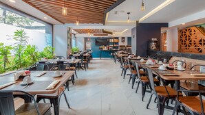 A modern cafe with wooden tables and chairs, a counter with a coffee machine, and a view of greenery outside.