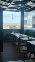 Un área de comedor moderna con mesas y sillas, vistas de la ciudad a través de amplias ventanas y un diseño de techo único.