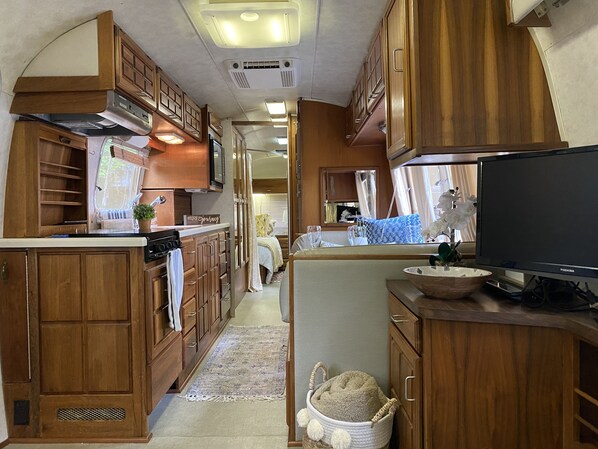 Cocina compacta para autocaravana con armarios de madera, estufa y televisor.