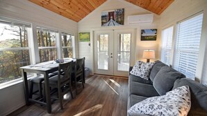 A cozy cabin interior with a dining area, a sofa, and large windows offering views of the surrounding forest. - Sweet Creek Retreat-Luxury Tiny Cabin-View-WIFi-Hot tub-Firepit Area-Smart TVs (Turtletown)