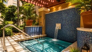 Una piscina con cascada, rodeada de plantas en macetas y un muro de azulejos.