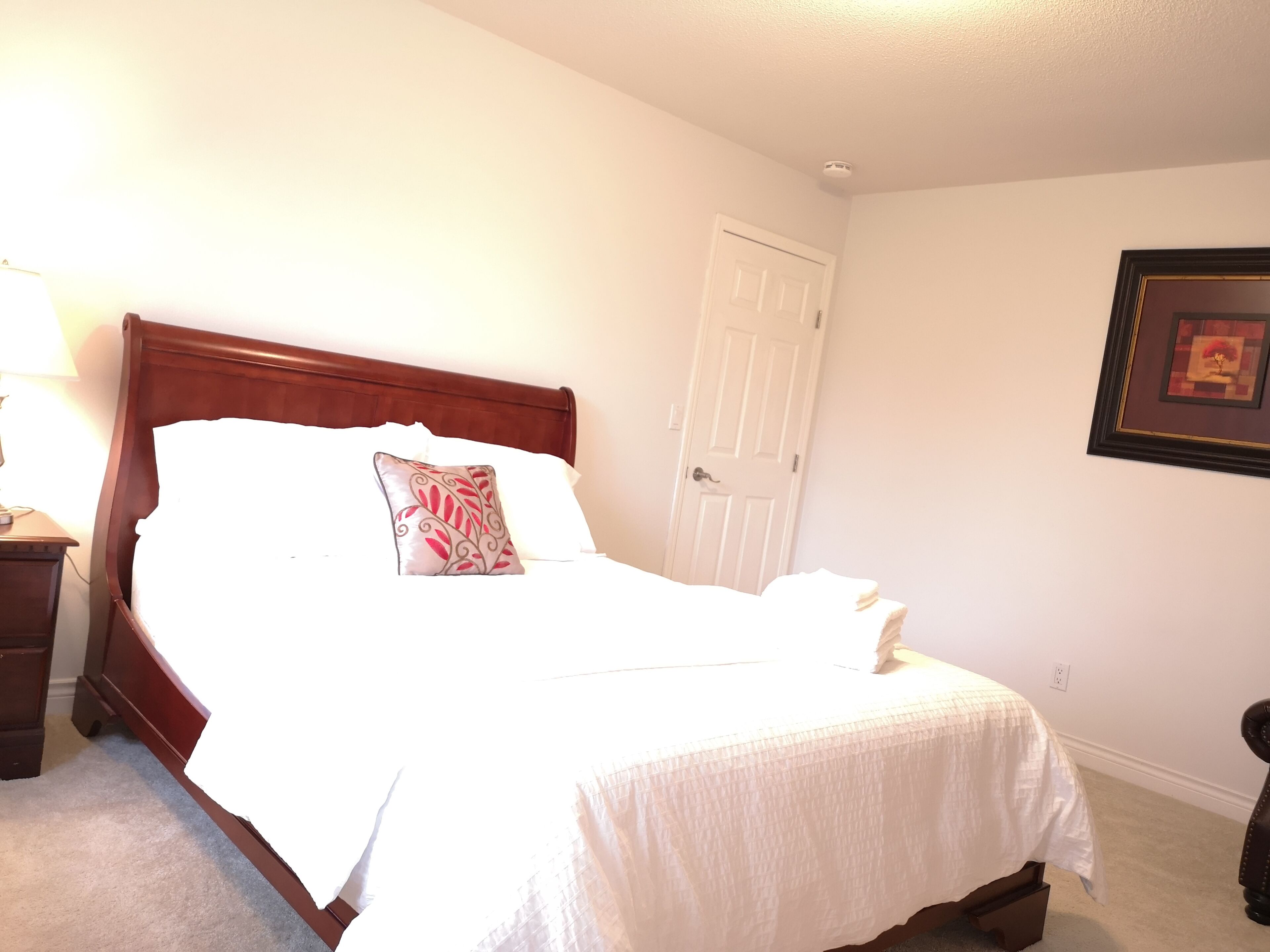 A neatly made bed with a white comforter and a decorative pillow, a wooden nightstand with a lamp, and a framed picture on the wall.